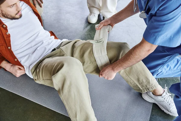 High angle view of professional paramedic applying compressive bandage on man in training room near interracial participants in first aid training seminar, bleeding prevention techniques concept — Stock Photo