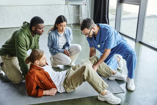 African american man and asian woman looking at medical instructor applying compressive bandage on leg of participant of first aid training seminar, bleeding prevention techniques concept — Stock Photo