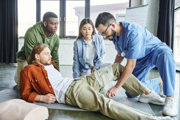 First aid seminar, medical instructor in eyeglasses and uniform applying compression bandage on leg of man near asian woman and african american, importance of emergency preparedness concept — Stock Photo