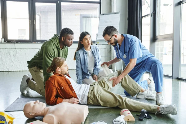 Medical training, professional instructor in uniform and eyeglasses bandaging leg of man near multicultural participants, CPR manikin and medical equipment, emergency preparedness concept — Stock Photo