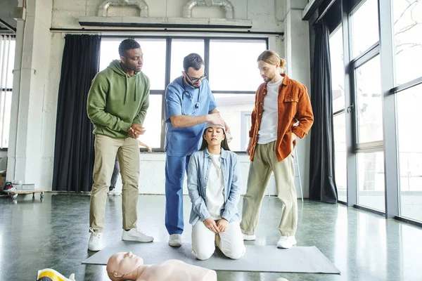 Multiethnischer Mann sieht während eines Erste-Hilfe-Trainings den professionellen Verbandskopf einer asiatischen Frau in der Nähe von CPR-Schaufensterpuppen an, Bedeutung des Notfallpräventionskonzeptes — Stockfoto