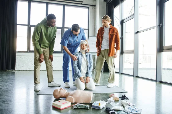 Hands-on first aid learning, young interracial men looking at healthcare worker bandaging head of asian woman near medical equipment and CPR manikin in training room, emergency response concept — Stock Photo