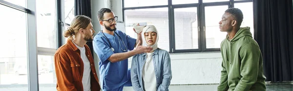 Smiling paramedic in eyeglasses bandaging head of young asian woman near multiethnic men while showing life-saving skills on first aid seminar, importance of emergency preparedness concept, banner — Stock Photo