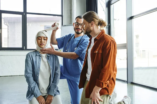 Positive medical instructor in eyeglasses and blue uniform looking at young man and bandaging head of asian woman during first aid seminar, important life-saving skills and techniques concept — Stock Photo
