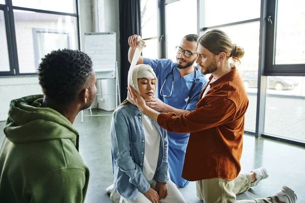 Joven hombre y médico instructor vendaje cabeza de asiático mujer cerca africano americano participante de primeros auxilios seminario en sala de formación, importancia de emergencia preparación concepto - foto de stock