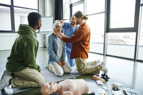 African american participant of first aid hands-on learning looking at young man and medical instructor bandaging head of asian woman near medical equipment, emergency preparedness concept — Stock Photo