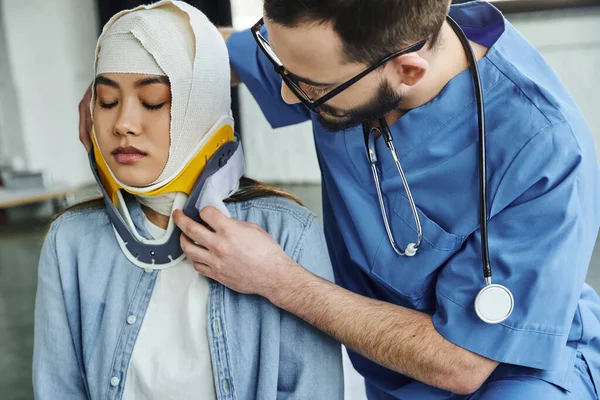 Paramédico profesional con estetoscopio, en uniforme y anteojos, poniendo soporte para el cuello en mujer asiática joven con cabeza vendada, entrenamiento médico, primeros auxilios y situaciones de emergencia concepto de respuesta - foto de stock