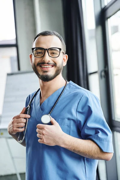 Überglücklicher bärtiger Ausbilder mit Stethoskop am Hals in blauer Uniform und lächelnd in die Kamera im Schulungsraum, Erste-Hilfe-Seminar und Notfallpräventionskonzept — Stockfoto
