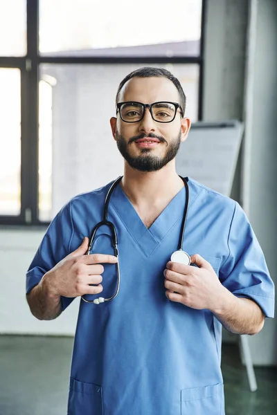 Fröhliche bärtige Gesundheitsfachangestellte mit strahlendem Lächeln in blauer Uniform und berührendem Stethoskop am Hals in Klinik, Erste-Hilfe-Schulung und Notfallpräventionskonzept — Stockfoto