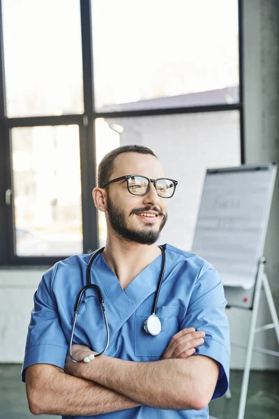 Happy bearded medical instructor in eyeglasses, blue uniform and stethoscope on neck crossing arms and looking away in training room, first aid training seminar and emergency preparedness concept — Stock Photo