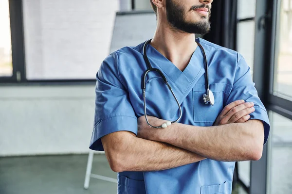 Teilaufnahme eines bärtigen Ausbilders in blauer Uniform, der mit verschränkten Armen und Stethoskop am Hals im Krankenhaus steht, Erste-Hilfe-Seminar und Notfallpräventionskonzept — Stockfoto