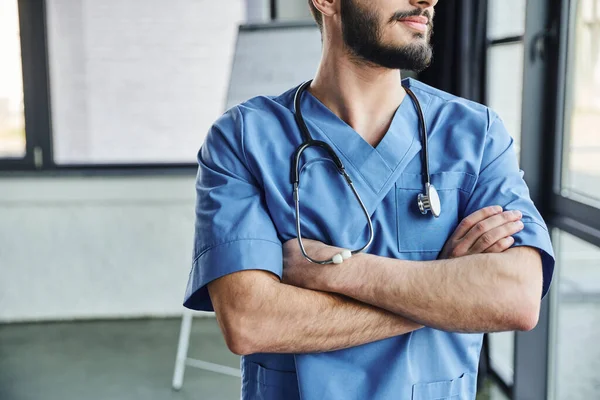 Vista parziale di istruttore medico barbuto professionista in uniforme blu in piedi con braccia incrociate e stetoscopio sul collo in ospedale, seminario di primo soccorso e concetto di preparazione alle emergenze — Foto stock