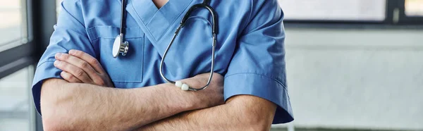 Vista parcial del joven médico en azul uniforme de pie con brazos cruzados y estetoscopio en el cuello en el hospital moderno, seminario de formación de primeros auxilios y concepto de preparación para emergencias, pancarta - foto de stock