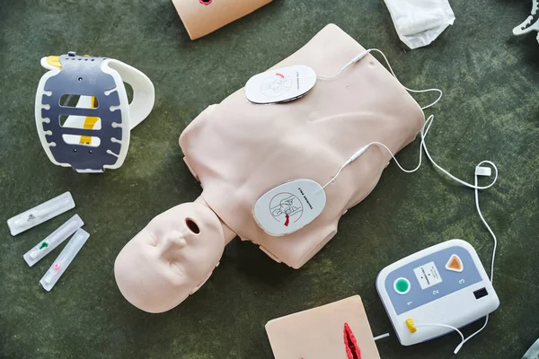 Top view of CPR manikin near automated defibrillator, wound care simulators, neck brace and syringes on floor in training room, medical equipment for first aid training and skills development — Stock Photo