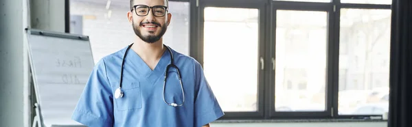 Happy bearded medical instructor in eyeglasses and blue uniform looking at camera while standing with stethoscope near flip chart on blurred background, first aid training seminar concept, banner — Stock Photo