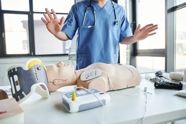 Cropped view of healthcare worker in blue uniform gesturing near CPR manikin with defibrillator near tourniquets and neck brace in training room, first aid hands-on learning concept — Stock Photo