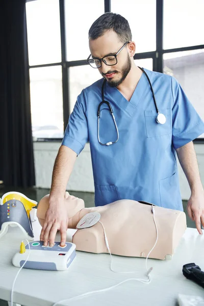 Healthcare worker in blue uniform, stethoscope and eyeglasses operating automated defibrillator near CPR manikin, first aid hands-on learning and critical skills development concept — Stock Photo