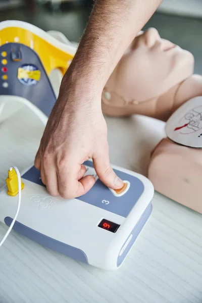 Teilansicht des Gesundheitshelfers, der den automatisierten Defibrillator bedient, während er die Herz-Wiederbelebung an einer CPR-Schaufensterpuppe übt, praktisches Erste-Hilfe-Lernen und ein Konzept zur Entwicklung kritischer Fähigkeiten — Stockfoto