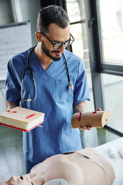 Bearded healthcare worker in eyeglasses and blue uniform, with stethoscope, holding wound care simulators above CPR manikin, first aid hands-on learning and critical skills development concept — Stock Photo