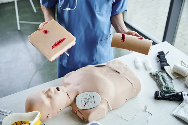Partial view of medical instructor standing with wound care simulator near CPR manikin with defibrillator and medical equipment, first aid hands-on learning and critical skills development concept — Stock Photo