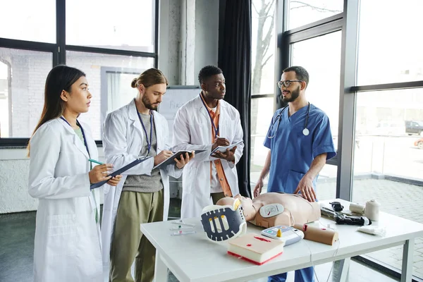 Professional paramedic talking to multiethnic students writing near CPR manikin, defibrillator and medical equipment during first aid seminar in training room, emergency situations response concept — Stock Photo