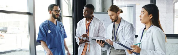 Gruppo giovane e diversificato di studenti multiculturali in camici bianchi che scrivono vicino all'istruttore medico parlando del seminario di primo soccorso in sala di formazione, concetto di risposta alle situazioni di emergenza, banner — Foto stock