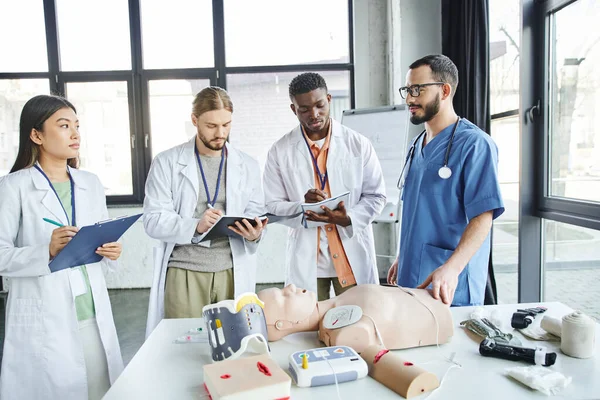 First aid seminar, diverse group of multiethnic students in white coats writing near paramedic, CPR manikin, defibrillator and medical equipment in training room, energy situations response concept — Stock Photo