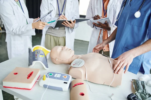 Cropped view of woman in white coat pointing at CPR manikin and defibrillator near paramedic and multiethnic students writing in notebooks, life-saving skills hands-on learning concept — Stock Photo