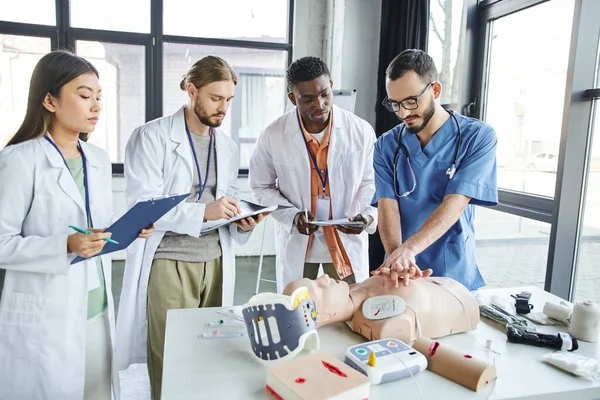 Medical instructor showing chest compressions on CPR manikin near defibrillator, medical equipment and multiethnic students writing in training room, emergency situations response concept — Stock Photo