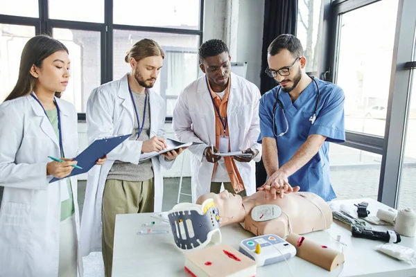 Instrutor médico mostrando compressões torácicas no manequim de RCP perto de desfibrilador, equipamentos médicos e estudantes multiétnicos escrevendo em sala de treinamento, conceito de resposta a situações de emergência — Fotografia de Stock