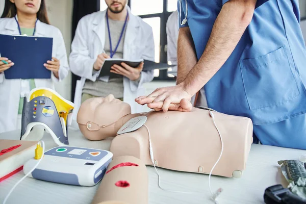 Partial view of paramedic practicing chest compressions on CPR manikin near defibrillator, neck brace and students with clipboard and notebook, life-saving skills hands-on learning concept — Stock Photo