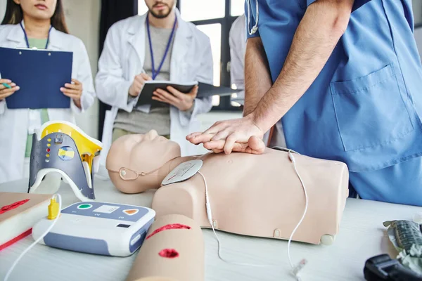 Vue partielle de l'ambulancier pratiquant des compressions thoraciques sur le mannequin CPR près du défibrillateur, de l'orthèse cervicale et des étudiants avec presse-papiers et bloc-notes, compétences salvatrices concept d'apprentissage pratique — Photo de stock