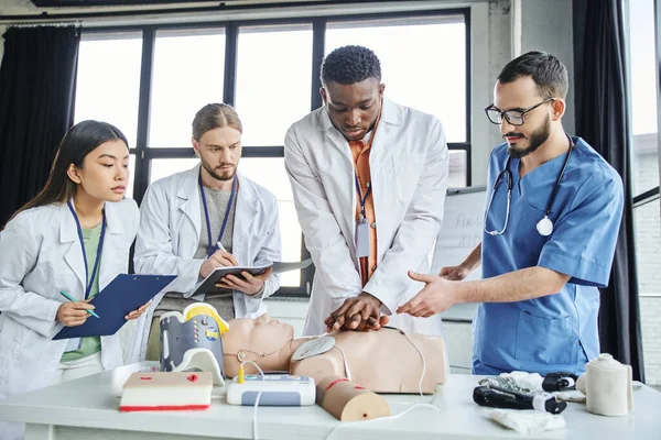 Homme afro-américain en blouse blanche pratiquant des compressions thoraciques sur mannequin CPR près des ambulanciers, du matériel médical et des étudiants multiethniques dans la salle d'entraînement, concept de réponse aux situations d'urgence — Photo de stock