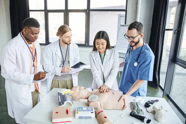 First aid training, cheerful asian woman practicing chest compressions on CPR manikin near medical instructor and multiethnic students writing in notebooks, emergency situations response concept — Stock Photo