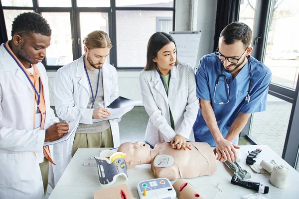 Multiethnic students in white coats writing in notebooks near medical instructor assisting asian woman doing chest compressions on CPR manikin, emergency situations response concept — Stock Photo