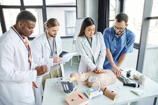First aid seminar, multiethnic students in white coats writing near asian woman doing chest compressions on CPR manikin with help of medical instructor, emergency situations response concept — Stock Photo