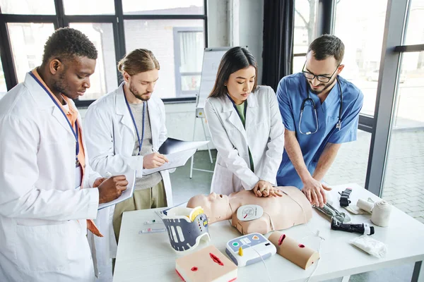 Seminario di primo soccorso, studenti multietnici in camici bianchi scrivere vicino a donna asiatica facendo compressioni toraciche sul manichino CPR con l'aiuto di istruttore medico, concetto di risposta alle situazioni di emergenza — Foto stock