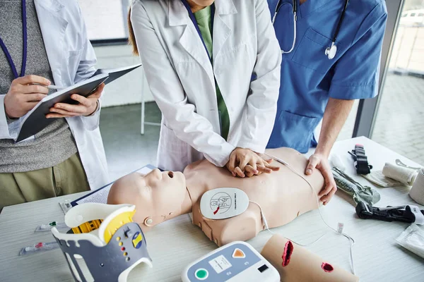 First aid seminar, partial view of young woman in white coat practicing chest compressions on CPR manikin near paramedic and student writing in notebook, life-saving skills hands-on learning concept — Stock Photo