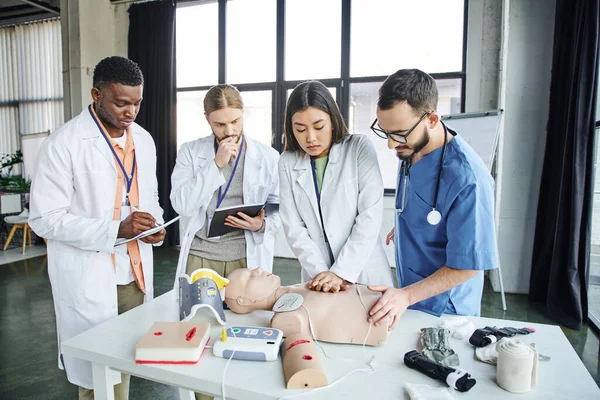 Young asian student in white coat practicing chest compressions on CPR manikin near defibrillator, healthcare worker, and multicultural men with notebooks, emergency situations response concept — Stock Photo
