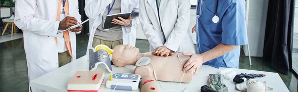 Partial view of woman in white coat doing chest compressions on CPR manikin near instructor while multiethnic students writing in notebooks, life-saving skills hands-on learning concept, banner — Stock Photo