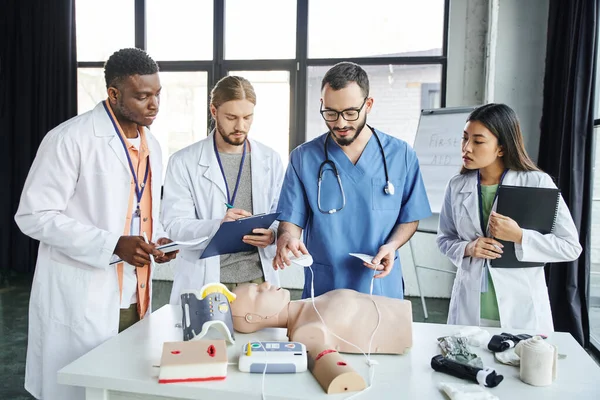 Cardiac resuscitation, medical instructor holding defibrillator pads above CPR manikin near young multiethnic students in white coats, emergency situations response concept — Stock Photo