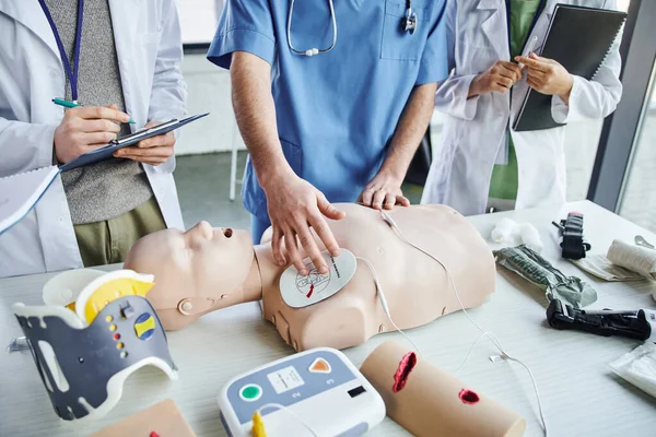 Vista recortada del instructor que aplica almohadillas de desfibrilador en el maniquí de RCP cerca del equipo médico y estudiantes jóvenes con batas blancas durante el seminario de primeros auxilios, habilidades que salvan vidas concepto de aprendizaje práctico - foto de stock