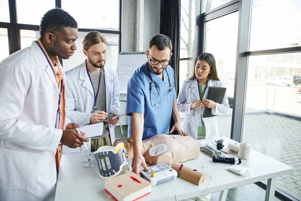 Healthcare worker showing defibrillator, CPR manikin and medical equipment to young multiethnic group in white coats standing with notebooks in training room, emergency situations response concept — Stock Photo
