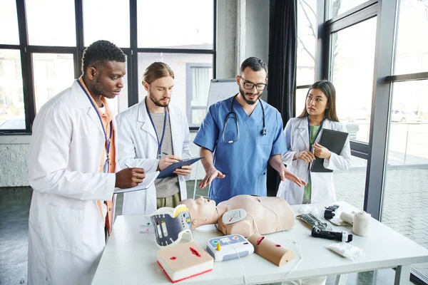 Professional paramedic talking and showing CPR manikin, external defibrillator and medical equipment to multiethnic students in white coats in training room, emergency situations response concept — Stock Photo
