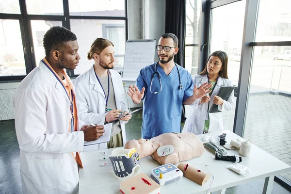 Professional paramedic talking to diverse group of multiethnic students near CPR manikin, automated defibrillator and medical equipment in training room, emergency situations response concept — Stock Photo