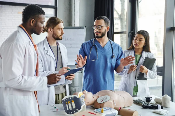 Multicultural students in white coats writing near healthcare worker gesturing and talking next to CPR manikin and medical equipment on first aid seminar, emergency situations response concept — Stock Photo