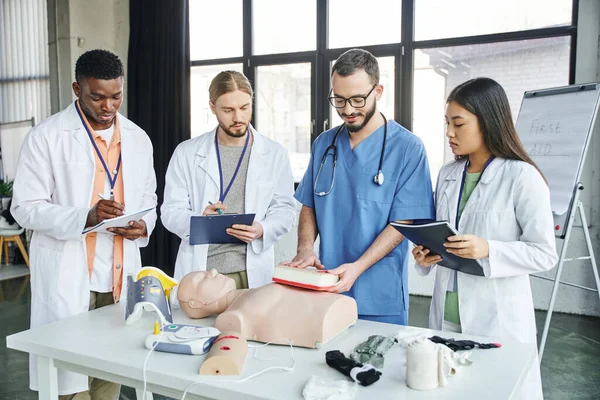 Multiethnic students writing next to instructor showing wound care simulator near CPR manikin, automated defibrillator and medical equipment in training room, emergency situations response concept — Stock Photo