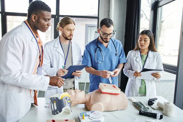 Healthcare worker holding bandage near wound care simulator and showing life-saving skills to multiethnic students in white coats standing with clipboards near medical equipment in training room — Stock Photo