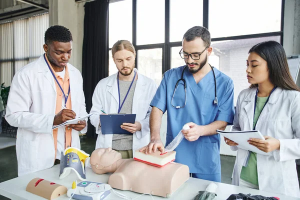 Multicultural students in white coats writing on clipboards and looking at medical instructor tamponing wound on simulator near CPR manikin and medical equipment, life-saving skills concept — Stock Photo