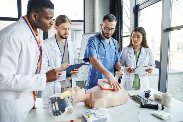 Professional paramedic tamponing wound on simulator near young multiethnic students in white coats and medical equipment during first aid training seminar, effective life-saving skills concept — Stock Photo